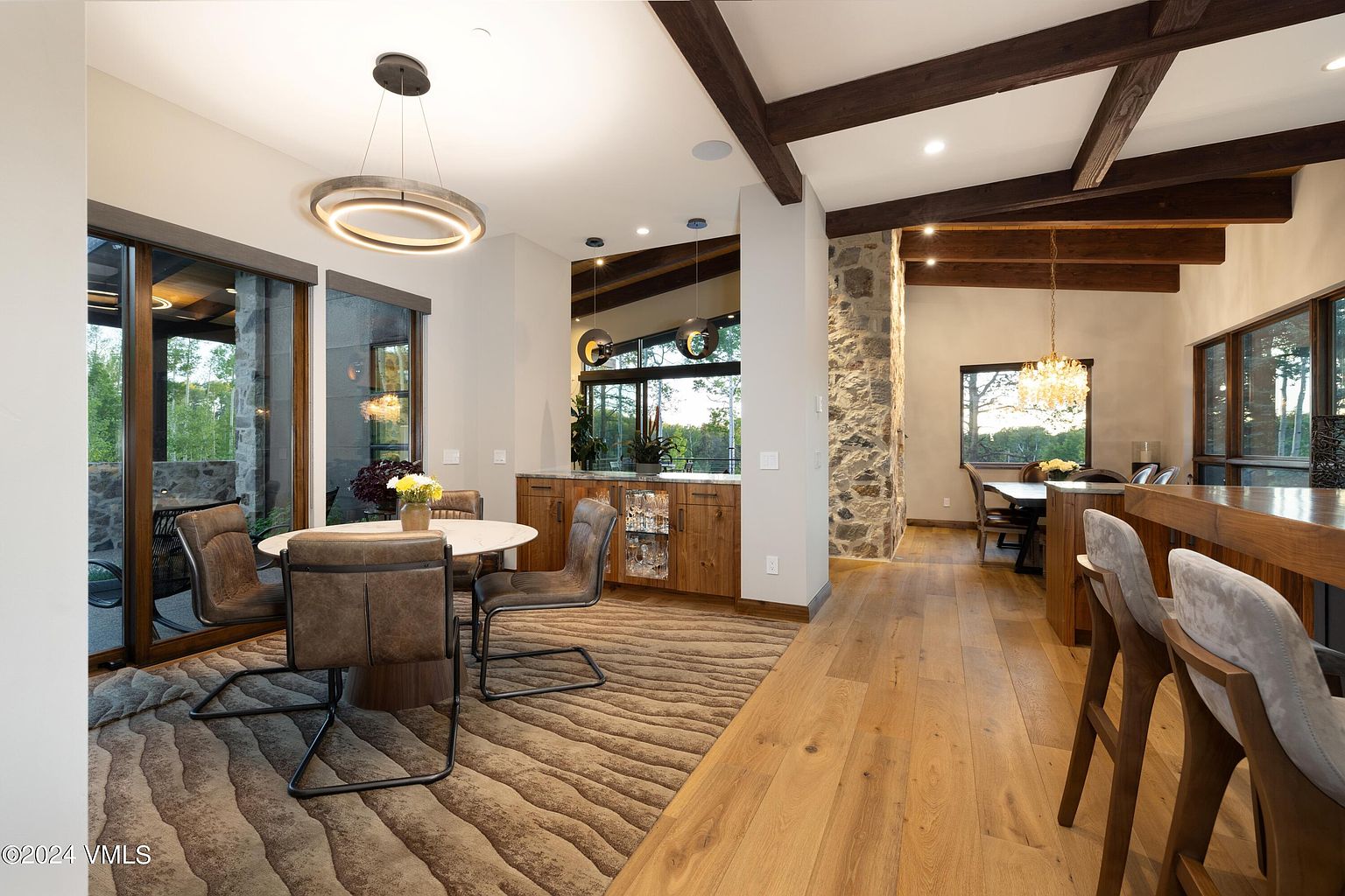 This interior shot showcases a dining area with a modern rustic design. A round table with four chairs sits atop a textured rug, illuminated by a circular pendant light. The space features wooden floors, exposed wooden beams, and a stone accent wall, creating a warm and inviting atmosphere.