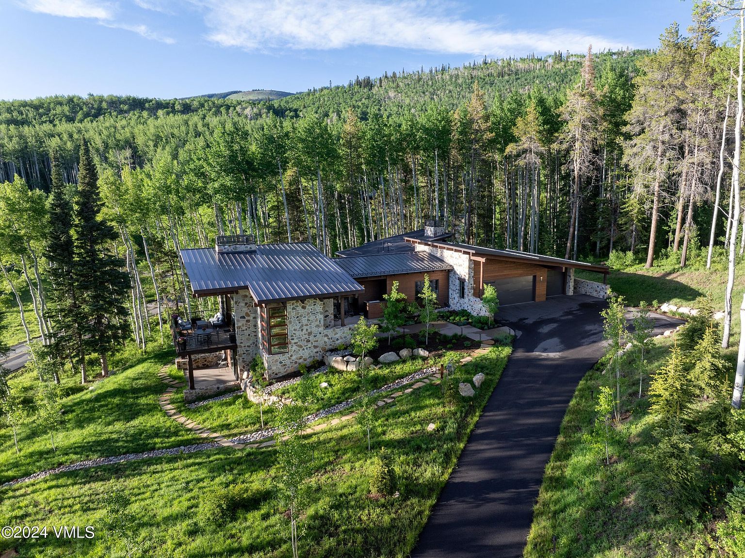 This aerial view showcases a stunning modern home nestled in a lush, forested landscape. The house features a stone facade, a metal roof, and a spacious deck, blending seamlessly with its natural surroundings. A paved driveway leads to an attached garage, and the meticulously maintained lawn and landscaping add to the property's curb appeal.