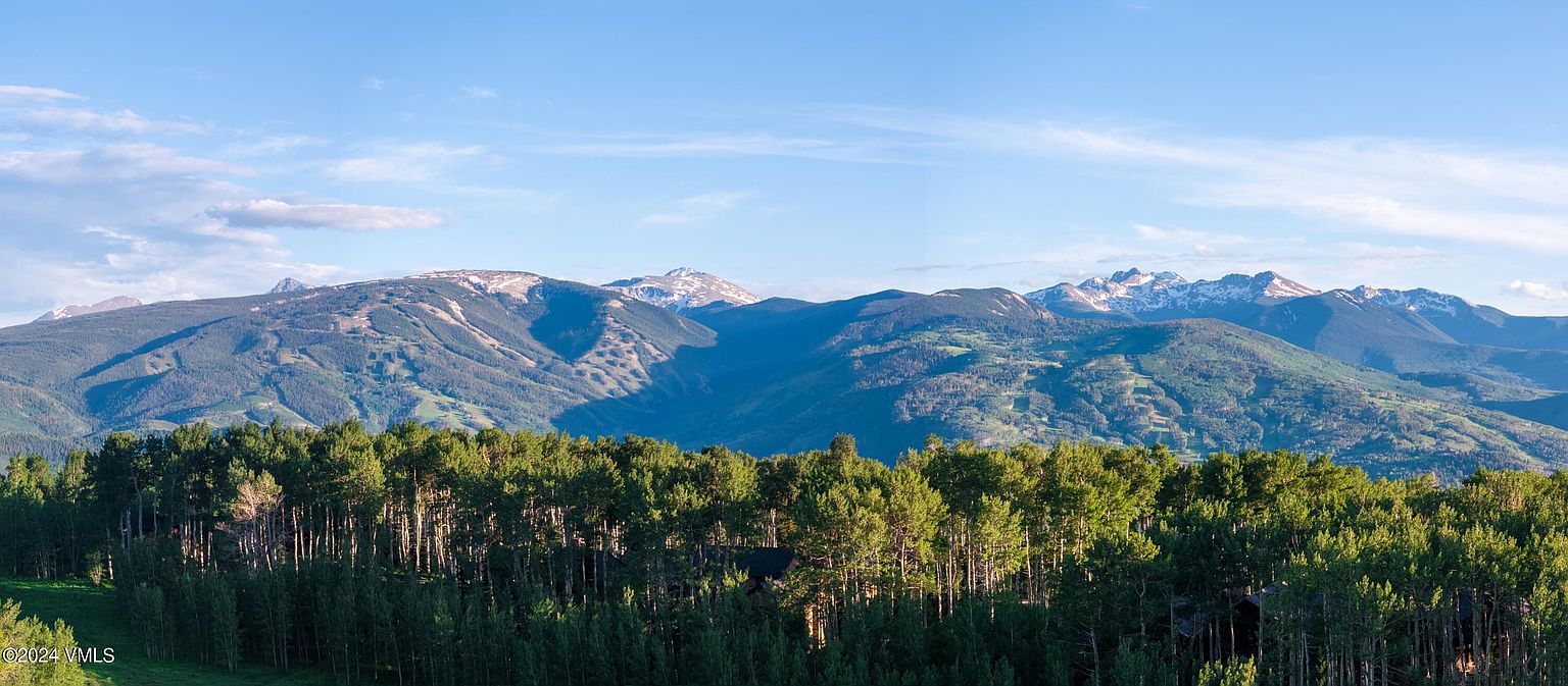 This stunning aerial view showcases a property nestled amidst a lush forest, with majestic mountains rising in the background. The landscape features a mix of evergreen and aspen trees, creating a vibrant tapestry of green and gold. The clear blue sky enhances the sense of tranquility and natural beauty, making this an ideal location for a serene retreat.