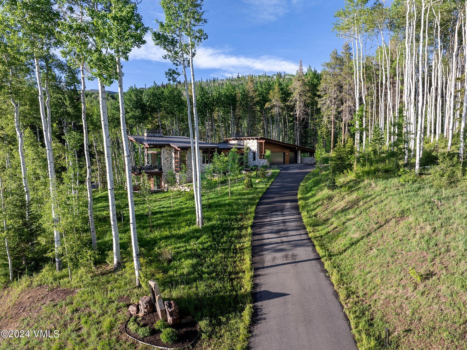 This image showcases the front exterior of a modern mountain home, nestled among aspen trees and lush greenery. A paved driveway leads up to the house, which features a stone facade and a contemporary roofline. The setting exudes privacy and tranquility, highlighting the property's natural surroundings and architectural design.