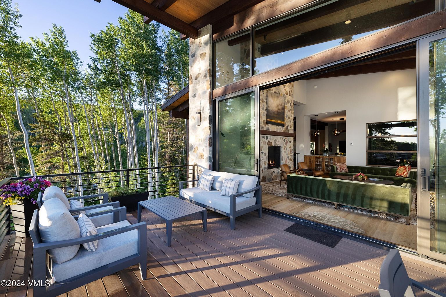 This image showcases a luxurious outdoor living space, featuring a spacious wooden deck with comfortable seating arrangements, including a sofa and armchairs. The deck seamlessly transitions into an open-concept living area through large sliding glass doors, offering stunning views of the surrounding forest. The stone accents on the building's exterior add a touch of rustic elegance to the modern design.
