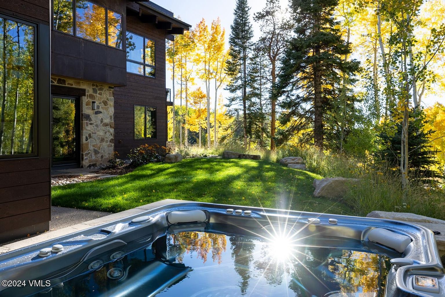 This image showcases a luxurious outdoor spa area, featuring a modern hot tub with crystal-clear water reflecting the surrounding trees and sunlight. The spa is situated next to a beautifully landscaped yard with lush green grass and mature trees, creating a serene and private retreat. The exterior of the house, with its stone and wood accents, adds to the upscale ambiance.