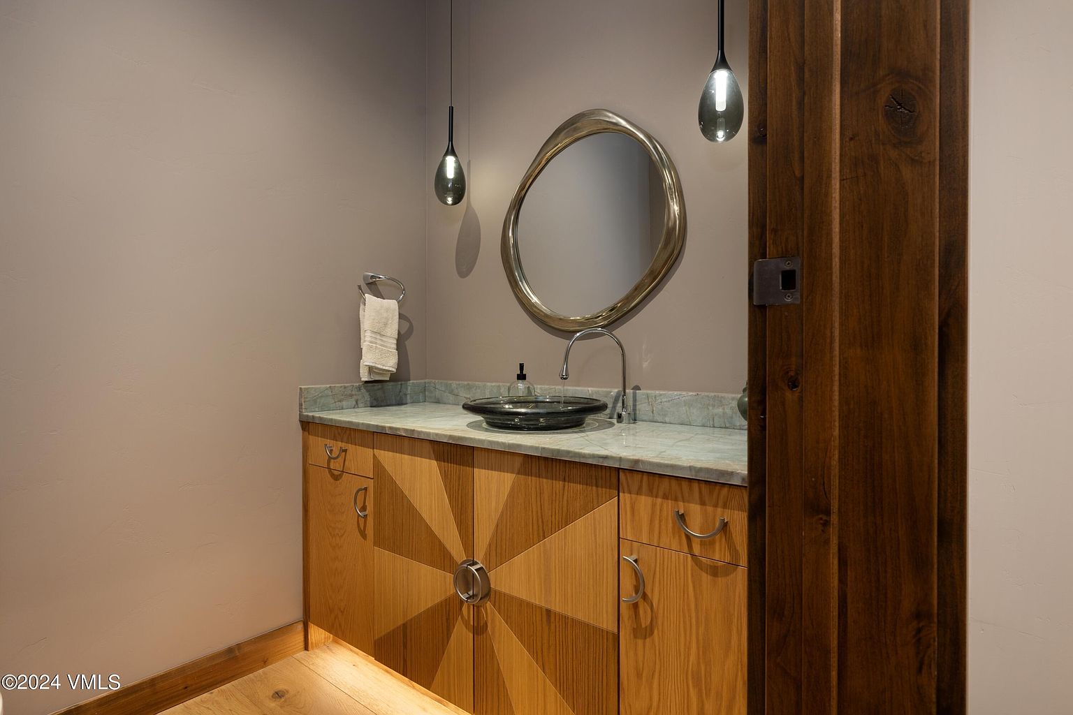 This is a well-appointed guest bathroom featuring a custom vanity with a unique starburst design and a light wood finish. The countertop is a light green marble, complemented by a modern, organically shaped mirror and pendant lighting. The overall aesthetic is sophisticated and contemporary, with a touch of rustic charm from the wooden door frame.