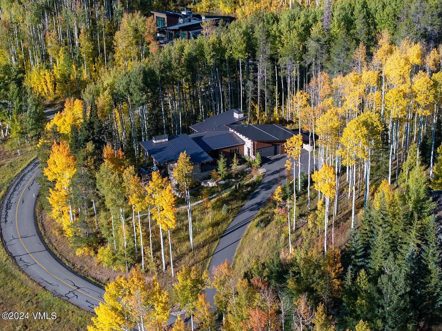 This aerial view showcases a modern home nestled among vibrant autumn foliage. The house features a dark roof, stone accents, and a winding driveway leading to a garage. The surrounding landscape is a mix of evergreen and deciduous trees displaying fall colors, creating a picturesque and secluded setting.