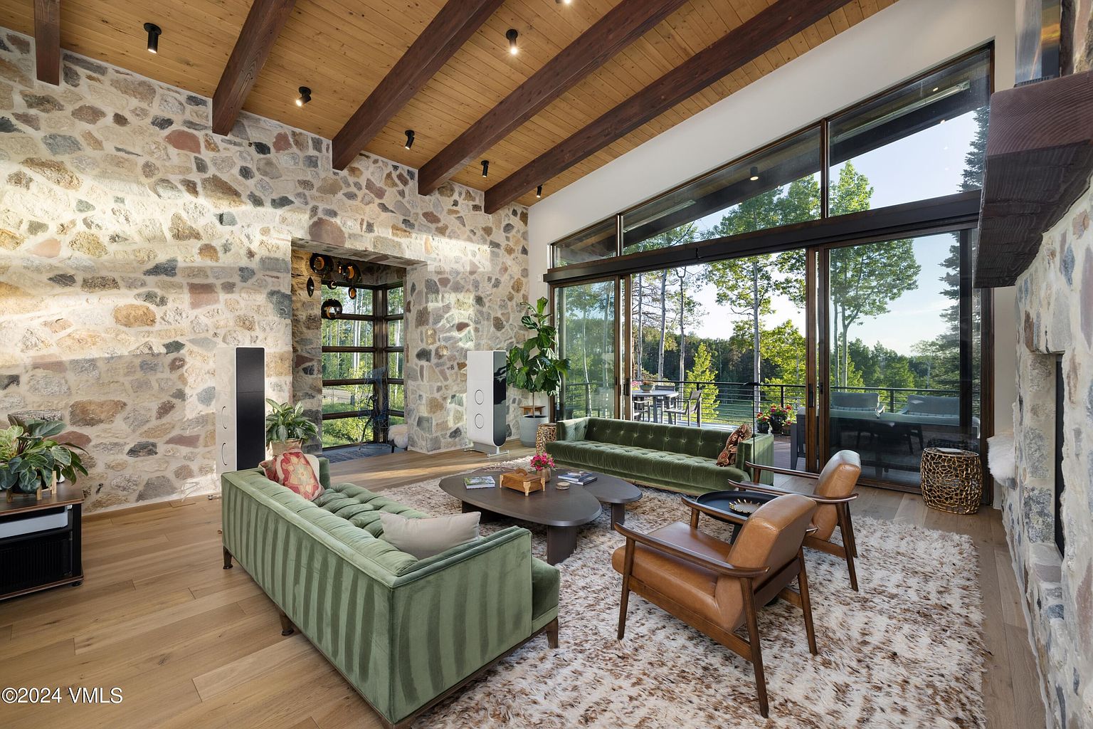 This is a spacious living room featuring a stone accent wall, wooden beamed ceiling, and large windows offering natural light and views of the outdoors. The room is furnished with green velvet sofas, leather chairs, and a patterned rug, creating a cozy and inviting atmosphere. The open layout and high ceilings contribute to a sense of grandeur and modern comfort.
