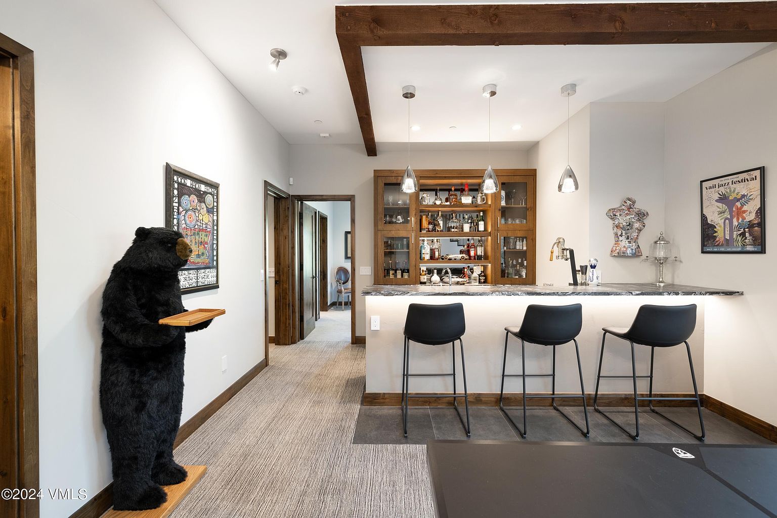 This interior shot showcases a stylish basement bar area, complete with a dark wood bar, glass-front cabinets stocked with beverages, and modern black bar stools. A decorative bear statue stands nearby, adding a unique touch to the space, while recessed lighting and pendant lights illuminate the area, creating a warm and inviting atmosphere.