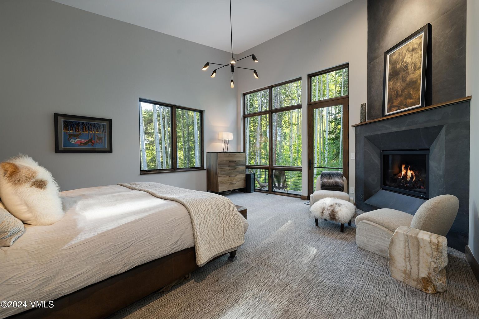 This primary bedroom features a modern design with large windows offering views of a forest. A stone fireplace adds warmth, complemented by neutral-toned furniture and a plush bed. The room is well-lit and spacious, creating a serene and luxurious atmosphere.