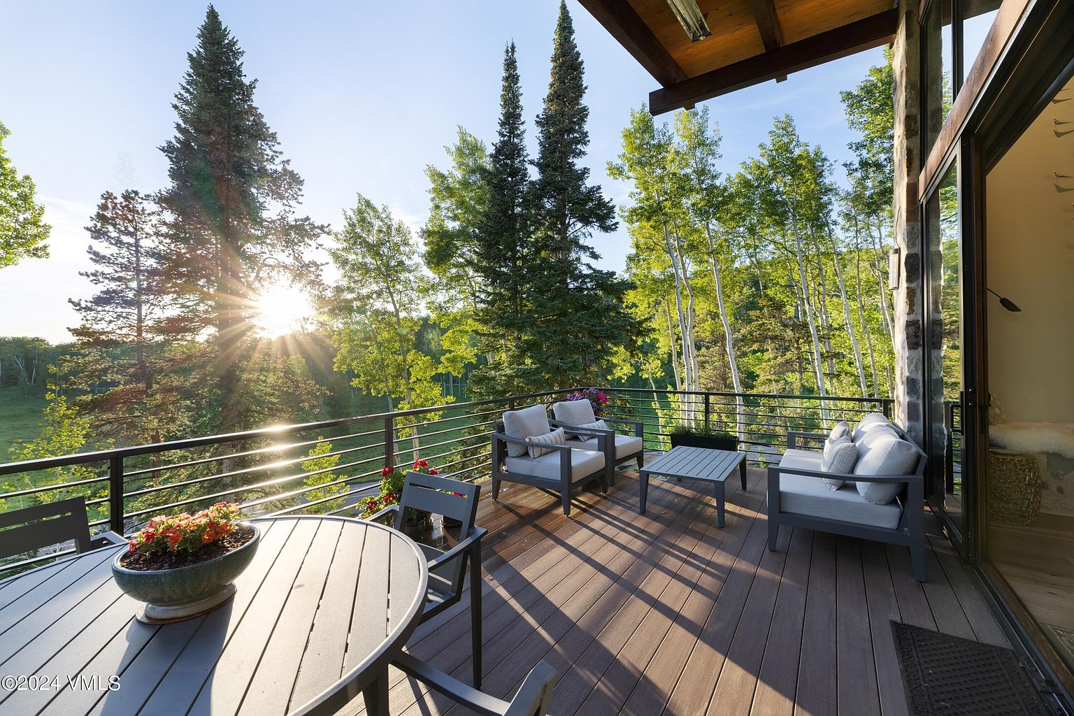 This image showcases a beautifully designed outdoor patio or deck area. The space features comfortable seating arrangements, including chairs and a table, perfect for relaxation or entertaining. The deck is surrounded by lush greenery and trees, offering a serene and private atmosphere. The sun shines through the trees, creating a warm and inviting ambiance.