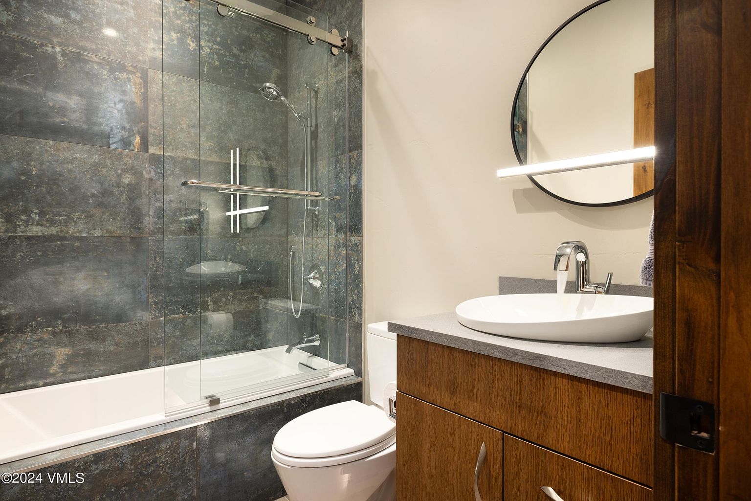 This is a well-appointed bathroom featuring a bathtub with a glass shower door, a modern toilet, and a vanity with a vessel sink. The walls are tiled with a dark, textured material, contrasting with the light-colored walls and white fixtures. A round mirror with a linear light fixture above it adds a touch of contemporary style.