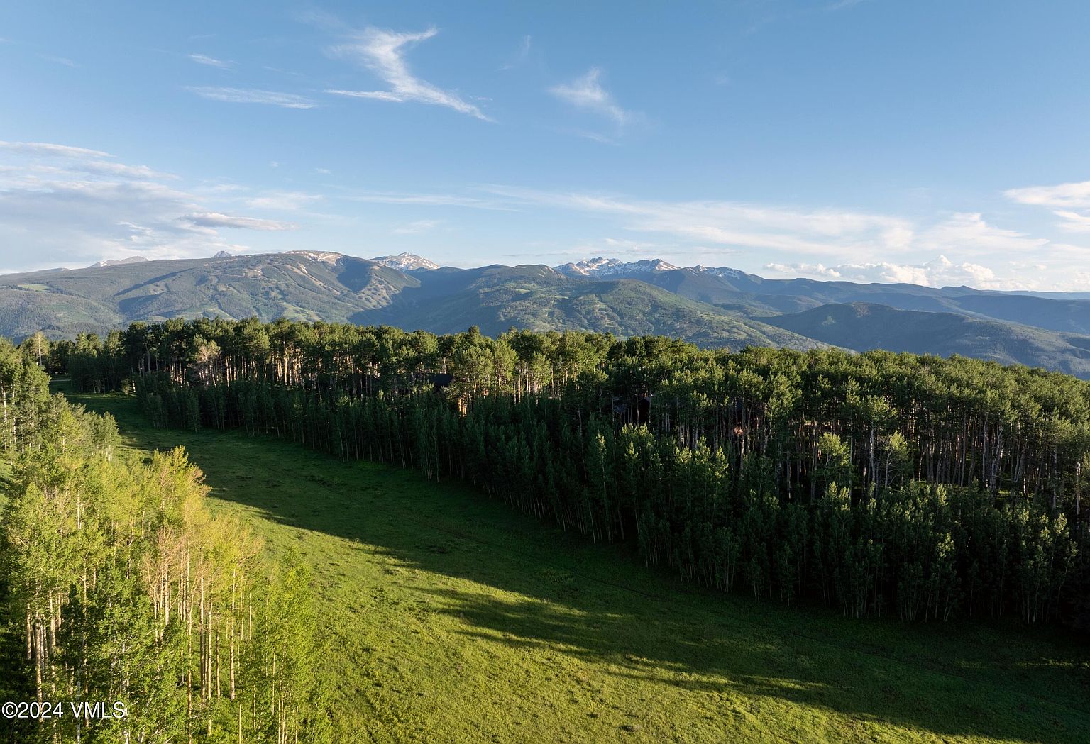 This aerial view showcases a lush green landscape with a dense forest in the foreground and majestic mountains in the background under a clear blue sky. The scene evokes a sense of tranquility and natural beauty, highlighting the property's serene setting and potential for outdoor recreation. The perspective emphasizes the vastness of the land and the stunning mountain views, making it an ideal location for a secluded retreat.
