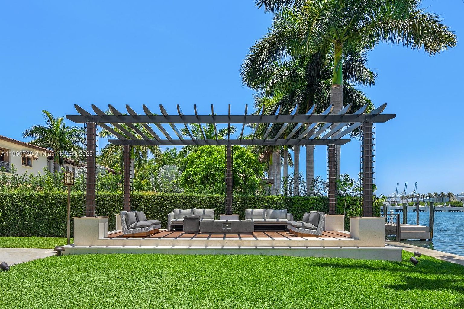 This elegant outdoor living space features a modern pergola structure covering a raised deck with comfortable, contemporary lounge seating. The area is set against a lush green hedge and tall palm trees, overlooking a serene waterfront with a view of distant industrial cranes. The scene captures a tranquil, upscale backyard retreat perfect for relaxation and entertaining.