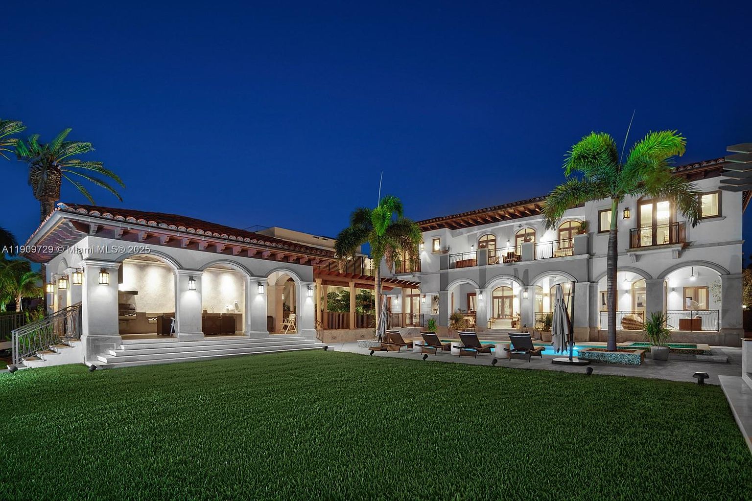 This stunning Mediterranean-style estate is captured at twilight, showcasing a sprawling backyard with a manicured lawn, a swimming pool, and an expansive covered outdoor kitchen area. The two-story main residence features elegant arched windows, balconies, and warm exterior lighting that highlights its architectural grandeur. The perspective is a wide, eye-level shot from the lawn, emphasizing the luxurious outdoor living space and the home's impressive scale.