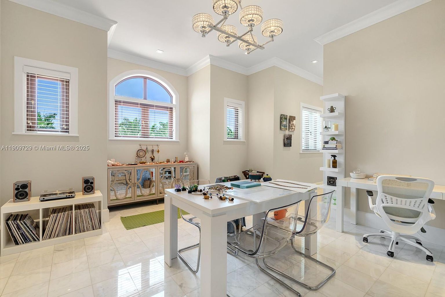 This bright and airy home office features a central white desk paired with modern transparent chairs, set against a backdrop of neutral walls and polished tile flooring. The room is accented by a sophisticated multi-bulb chandelier, a mirrored sideboard, and a tall white shelving unit, creating a clean, contemporary workspace. A large arched window allows for ample natural light, enhancing the open and inviting atmosphere of the room.