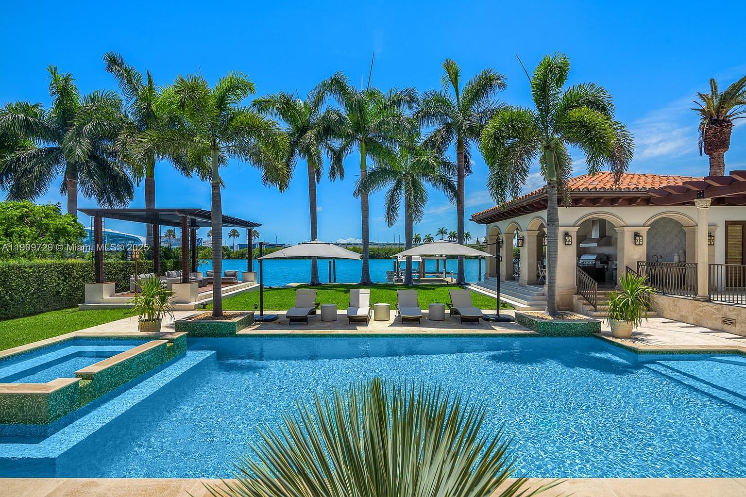 This stunning waterfront property features a pristine, turquoise swimming pool in the foreground, leading the eye toward a lush green lawn and a row of elegant palm trees. The scene includes a covered patio area with lounge chairs and a view of the sparkling blue bay, creating a luxurious and serene tropical atmosphere. The perspective is captured from a low angle at the pool's edge, emphasizing the seamless transition between the home's outdoor living space and the water.