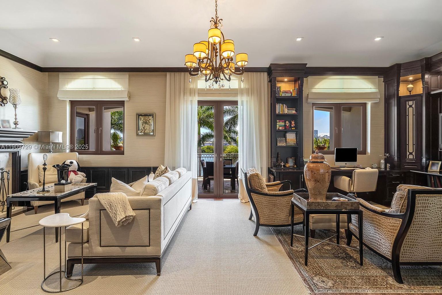 This sophisticated home office and living space features a blend of classic elegance and functional design, highlighted by dark wood built-in cabinetry and a central chandelier. The room is furnished with a plush cream sofa, patterned armchairs, and a dedicated workspace with a computer, all arranged to overlook a balcony through French doors. The neutral color palette, textured wallpaper, and ornate rug create a refined, professional, and comfortable atmosphere.