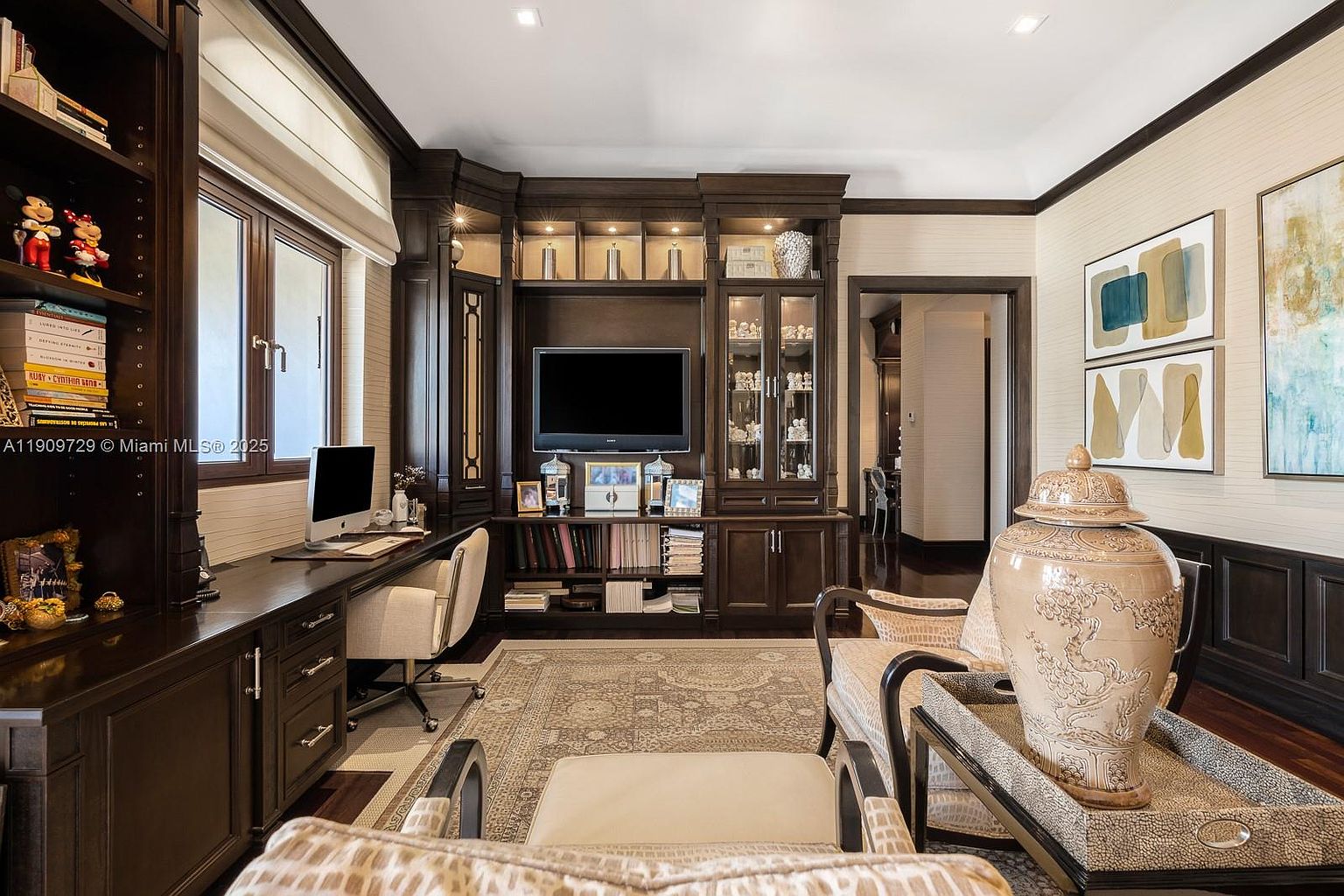 This sophisticated home office features rich, dark wood built-in cabinetry, a spacious desk area, and elegant decorative accents. The room is anchored by a patterned area rug and comfortable seating, creating a professional yet inviting atmosphere. The perspective is from the center of the room, looking toward the desk and media center, highlighting the high-end finishes and organized layout.