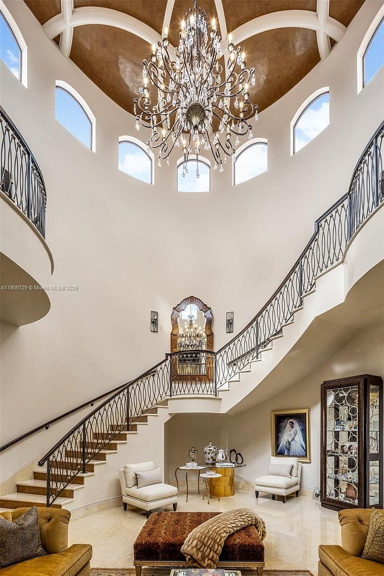 This grand foyer features a dramatic double-curved staircase with intricate wrought-iron railings, leading up to a soaring, multi-story rotunda ceiling. A magnificent crystal chandelier serves as the centerpiece, hanging above a luxurious seating area with plush armchairs and a central ottoman. The space is finished with polished marble flooring and elegant architectural details, creating an atmosphere of opulence and sophistication.