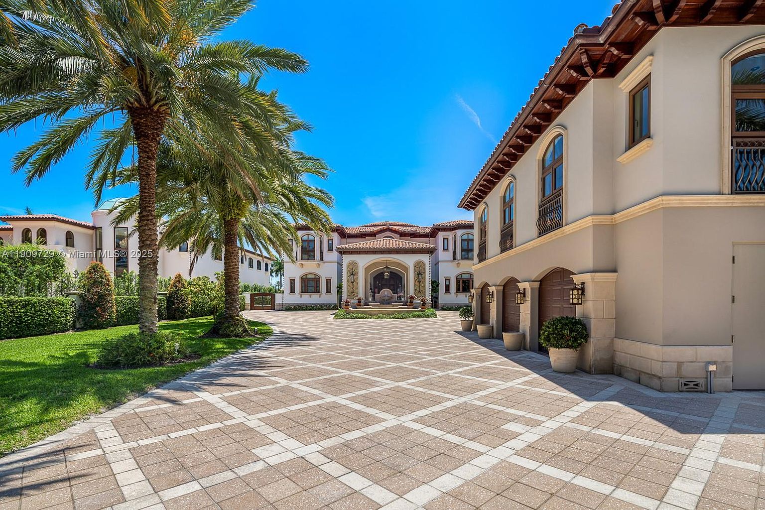 This grand Mediterranean-style estate features an expansive, meticulously paved motor court leading to a multi-car garage and a stately main entrance. The property is framed by lush landscaping, including a prominent palm tree, and showcases elegant architectural details like arched windows and a tiled roof. The perspective is a wide-angle shot from the driveway, emphasizing the scale and luxury of the residence under a clear blue sky.