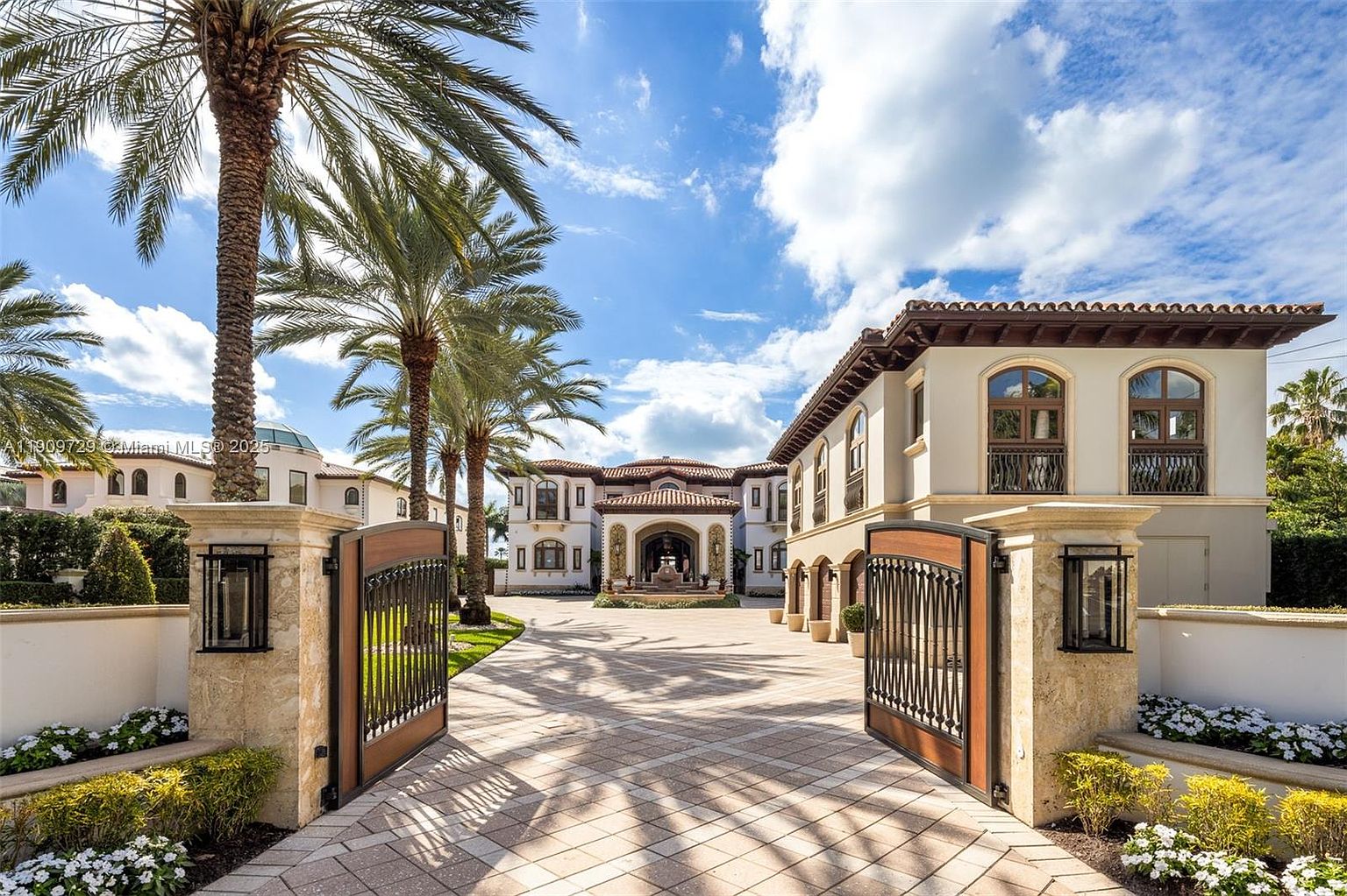 This grand Mediterranean-style estate features an expansive paved motor court framed by ornate iron gates and lush palm trees. The symmetrical architecture showcases elegant arched windows, a tiled roof, and a sophisticated entrance, creating an impression of luxury and exclusivity. The bright, sunny sky enhances the warm tones of the building's facade, highlighting its impressive curb appeal.