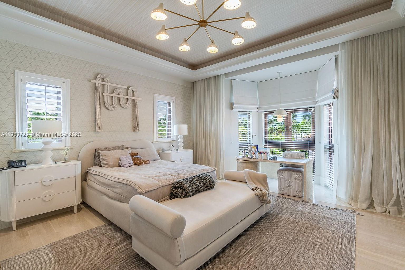 This elegant primary bedroom features a neutral color palette with soft textures, a plush upholstered bed, and a matching bench at the foot. A modern gold chandelier serves as a focal point, while a dedicated workspace is tucked into the sunlit bay window area. The room exudes a serene and sophisticated atmosphere, enhanced by layered window treatments and patterned wallpaper.