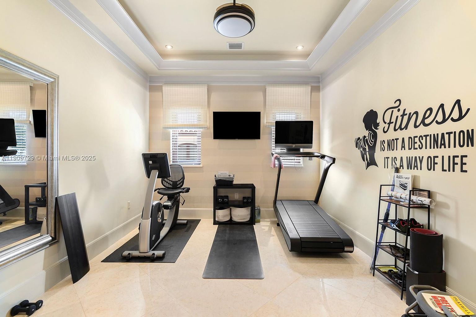 This well-lit home gym features a neutral color palette with a large wall mirror on the left and an inspirational wall decal on the right. The space is equipped with a stationary bike, a treadmill, and a central storage unit, all set upon a light-colored tile floor. The room is symmetrically arranged with two windows covered by blinds, creating a clean and focused environment for exercise.