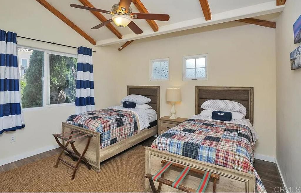This is a cozy guest bedroom featuring two twin beds with matching plaid quilts and wooden headboards. Natural light streams in through a window adorned with blue and white striped curtains. The room has a warm and inviting atmosphere, enhanced by the wooden ceiling beams and neutral wall color.