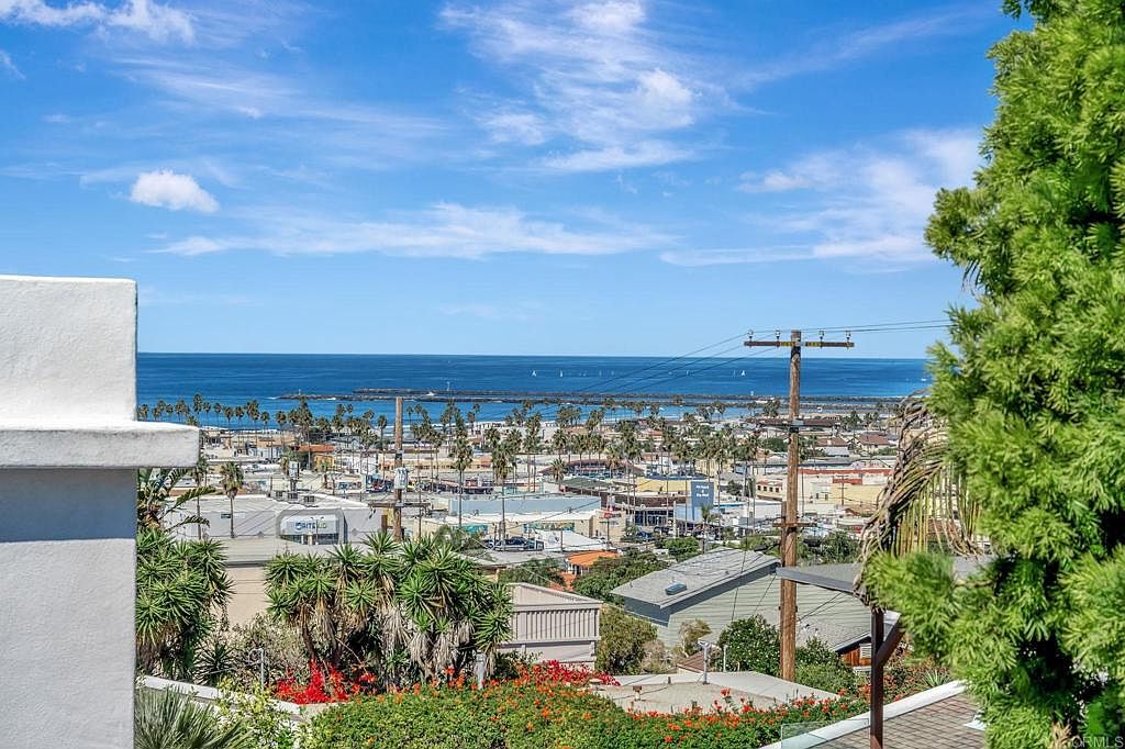 This aerial view showcases a coastal town with a clear blue ocean backdrop. Palm trees line the streets, and various buildings are visible, suggesting a mix of residential and commercial properties. The image captures a sunny day, highlighting the town's proximity to the ocean and its overall pleasant atmosphere.