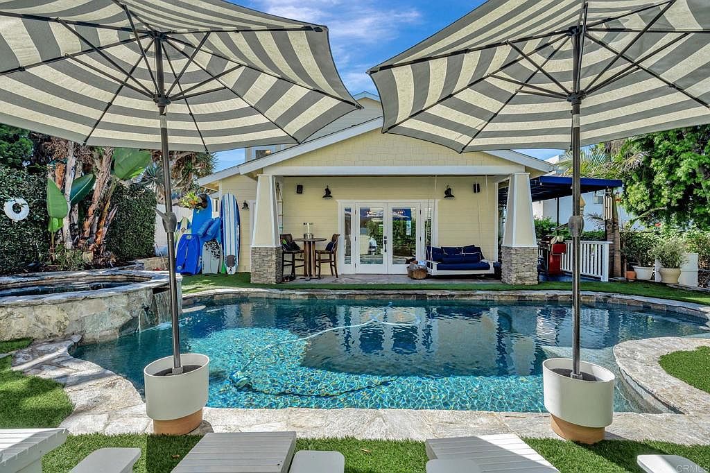 This image showcases a beautiful backyard pool area, featuring a freeform pool with a stone waterfall feature and lush landscaping. Two large striped umbrellas provide shade over lounge chairs, creating a relaxing atmosphere. The house, with its yellow siding and French doors, serves as a charming backdrop to the pool area, enhancing the property's appeal.