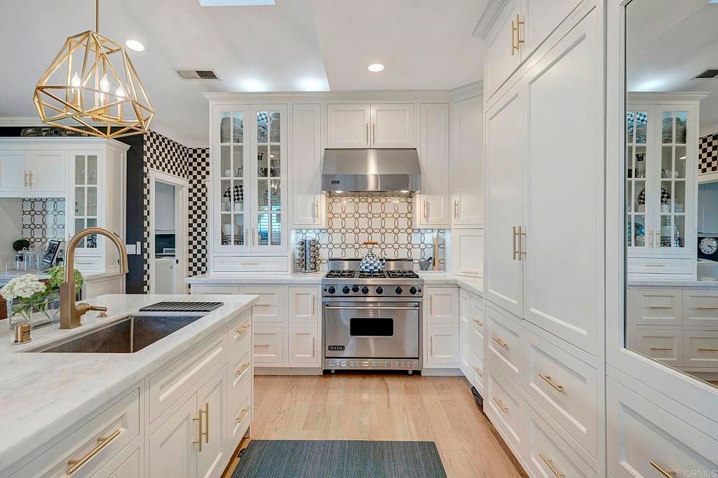This is a bright and elegant kitchen featuring white cabinetry with gold hardware, stainless steel appliances, and a marble countertop island. The kitchen is well-lit with a modern gold chandelier and recessed lighting, and the backsplash behind the stove adds a decorative touch. The overall impression is one of luxury and sophistication.