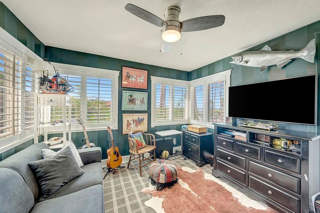 This is a cozy bedroom featuring a blue-gray sofa, a dark wood dresser with a large television, and a mounted fish decoration. The room has a nautical or rustic theme, with striped wallpaper, plantation shutters, and a plaid rug. Natural light floods the room through the windows, creating a warm and inviting atmosphere.