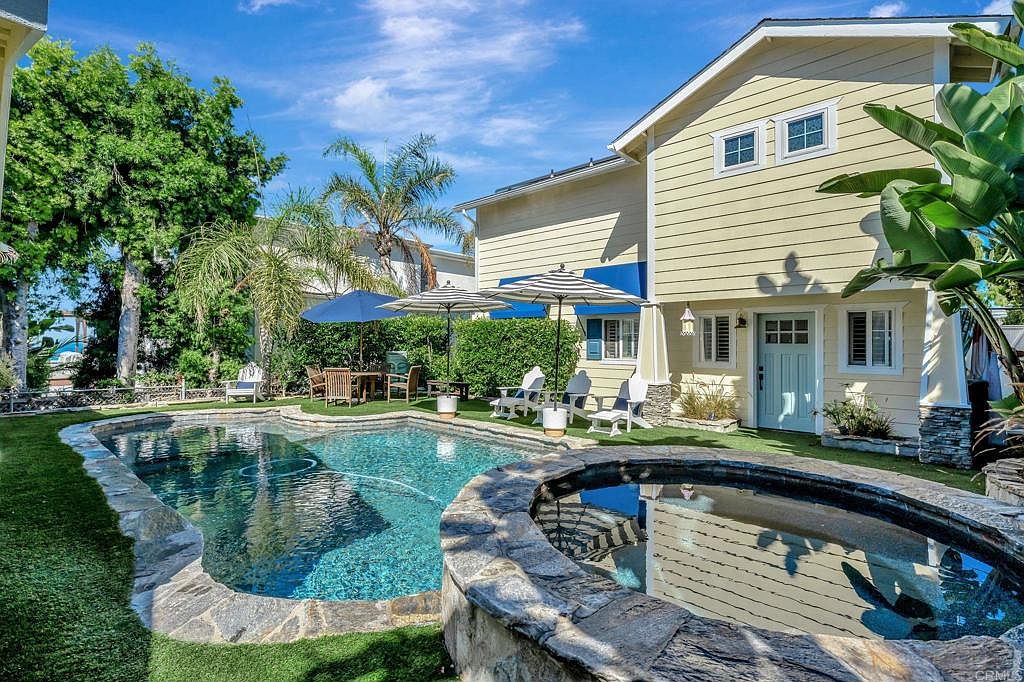 This image showcases a beautifully landscaped backyard featuring a kidney-shaped pool and a circular spa, both bordered by natural stone. The pool's clear water reflects the surrounding greenery and the house, which has a light yellow exterior with a charming blue door. The scene is framed by lush trees and manicured grass, creating a serene and inviting outdoor living space.