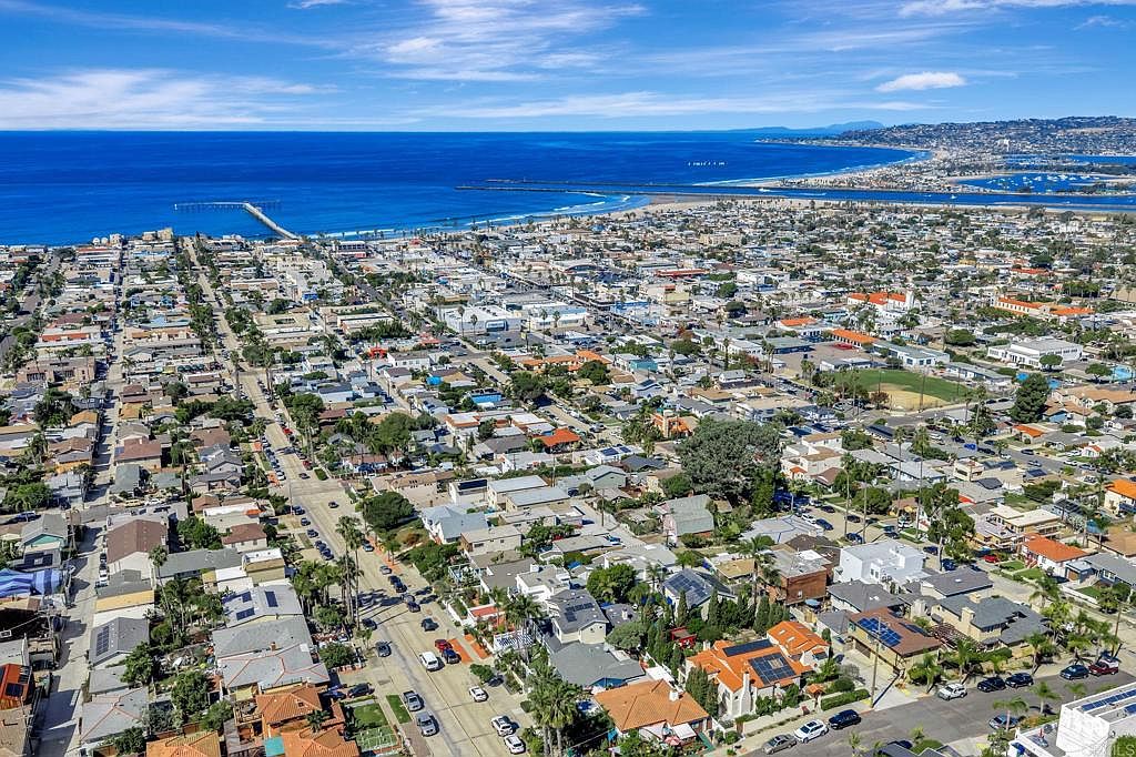 This aerial view showcases a dense residential neighborhood near the ocean. The houses are a mix of styles, many with solar panels, and are interspersed with mature trees. The scene captures the proximity to the beach and the overall coastal community vibe.