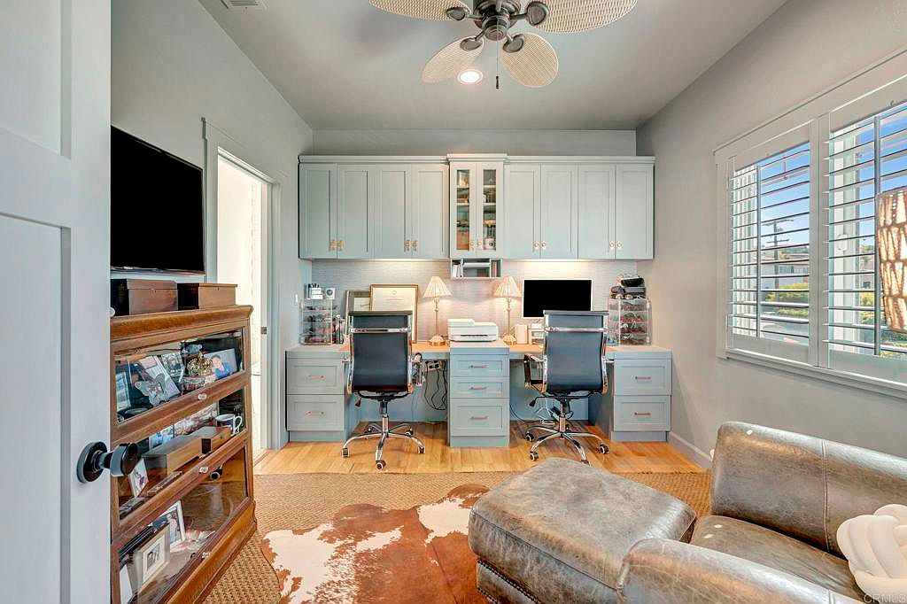 This is an interior shot of a well-organized home office featuring built-in cabinetry and dual workstations. The room is decorated in a neutral color palette with light blue cabinets and wooden accents. A comfortable seating area with a leather chair and ottoman adds a touch of relaxation to the workspace.