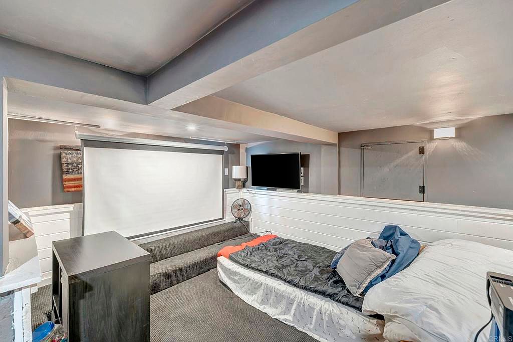 This is a cozy bedroom featuring a bed with a gray and orange comforter, a white headboard, and several pillows. A large projector screen is positioned on one wall, and a flat-screen TV is mounted on the adjacent wall. The room has gray walls and a carpeted floor, creating a comfortable and inviting space.