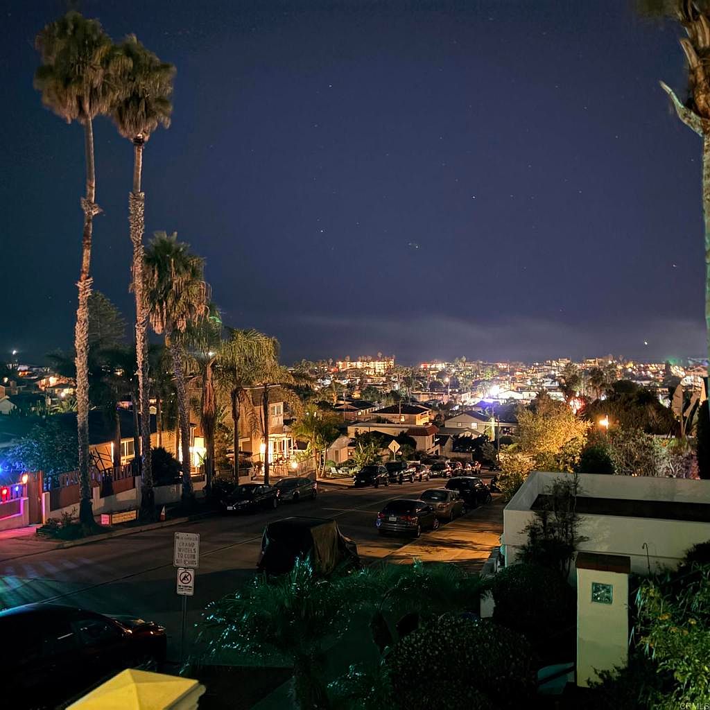 This aerial view captures a charming neighborhood at night, with twinkling city lights in the distance. Palm trees frame the scene, adding a touch of tropical elegance. The well-lit streets and cozy homes create a warm and inviting atmosphere, perfect for showcasing the community's appeal.