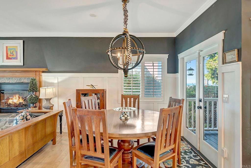 This is an interior shot of a dining room featuring a round wooden table with six chairs. A unique spherical chandelier hangs above the table, and natural light streams in through a window and glass doors leading to a balcony. The room is decorated in a classic style with dark gray walls and white wainscoting, creating an elegant and inviting atmosphere.