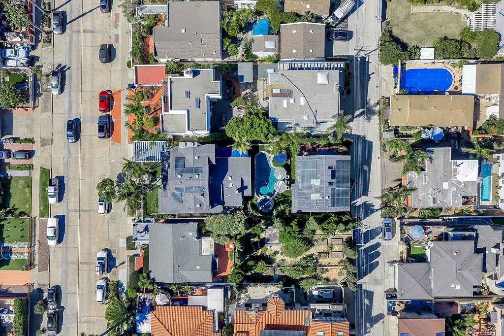 This aerial view showcases a residential neighborhood with well-maintained homes, lush landscaping, and several swimming pools. The houses feature varying roof styles, some with solar panels, and are situated along a tree-lined street with parked cars. The overall impression is one of a desirable and established community.