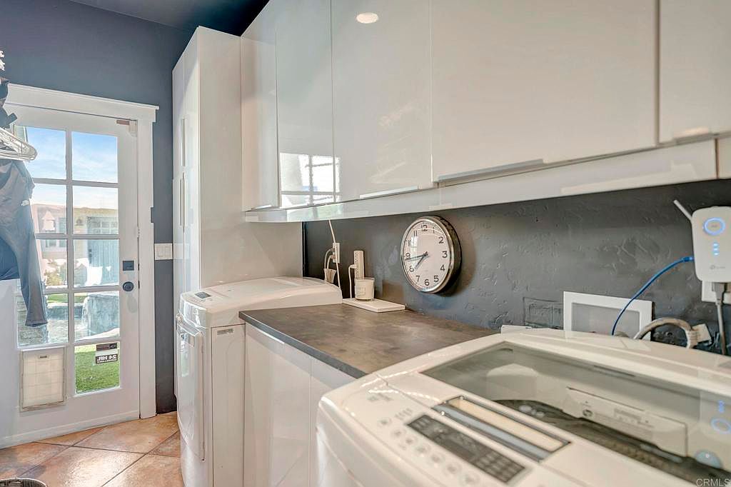 This is an interior shot of a modern laundry room. The room features white cabinetry, a dark countertop, and a front-loading washer and dryer. A round clock hangs on the wall above the countertop, and a door with glass panes leads to the outside.