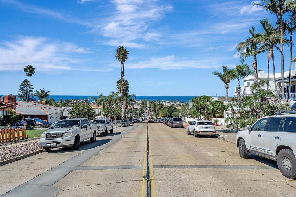 This image showcases a street view with a slight downward slope towards the ocean in the distance. Several cars are parked along the sides of the road, and palm trees line the street, adding a tropical feel. The houses on either side of the street appear well-maintained, contributing to an overall impression of a desirable residential neighborhood.