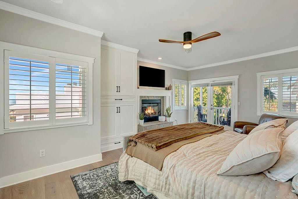 This is a primary bedroom featuring a fireplace with a mounted television above it, built-in cabinets, and a large window with shutters. The room has a neutral color palette, hardwood floors, and a ceiling fan. Sliding glass doors lead to a balcony, enhancing the room's appeal.