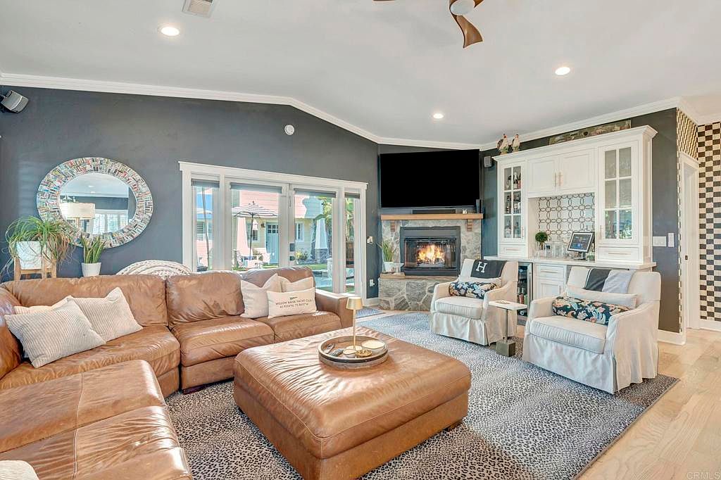 This is an interior shot of a living room featuring a large brown leather sectional sofa and ottoman, a stone fireplace with a mounted television, and two armchairs flanking a built-in bar area. The room is decorated with a patterned rug and a round mirror, creating a cozy and inviting atmosphere. The perspective is from the center of the room, capturing the main features.