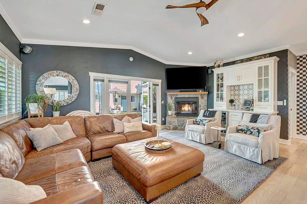 This is a warm and inviting living room featuring a large, comfortable leather sectional sofa and a matching ottoman. The room is centered around a stone fireplace with a mounted television above, flanked by built-in cabinetry. A patterned rug anchors the space, and natural light streams in through the windows, creating a cozy atmosphere.
