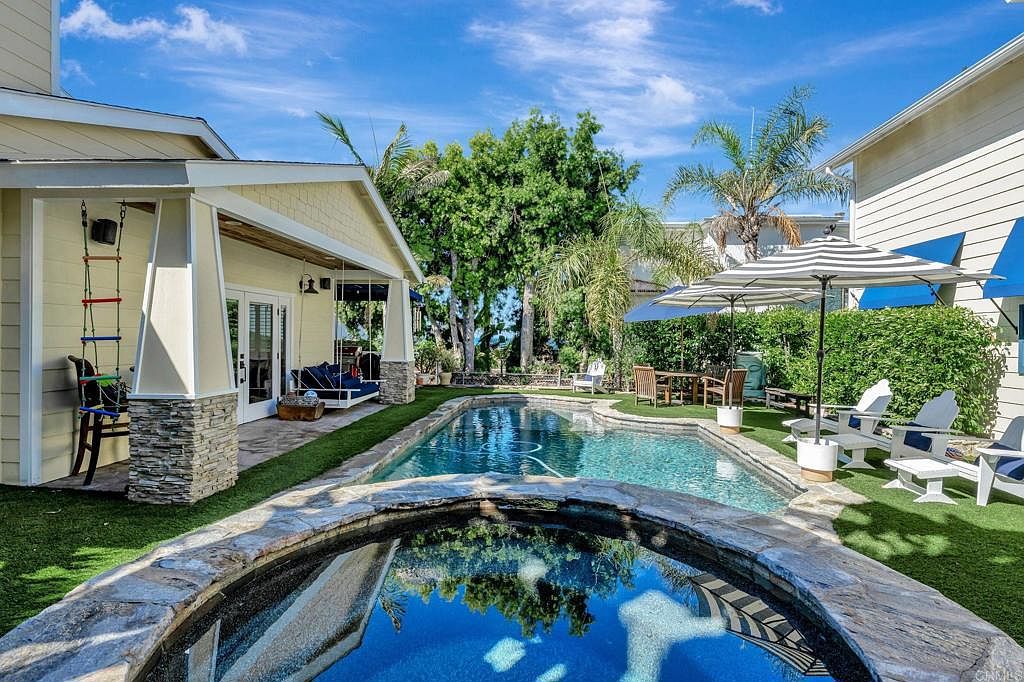 This image showcases a luxurious backyard pool and spa area. The pool is surrounded by well-maintained artificial turf and stone coping, with lounge chairs and umbrellas providing a relaxing atmosphere. The spa is in the foreground, reflecting the sky and surrounding trees, creating a serene and inviting outdoor space.