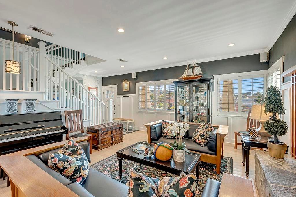 This is a warm and inviting living room featuring hardwood floors, a dark accent wall, and a staircase. The room is furnished with comfortable sofas adorned with floral pillows, a piano, and a decorative cabinet displaying nautical items. Natural light streams in through shuttered windows, creating a cozy and elegant atmosphere.
