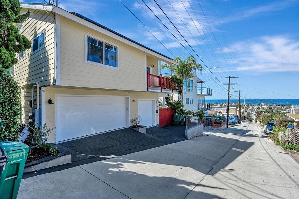 This is a front exterior view of a two-story house with a light yellow facade. The house features a two-car garage with white doors, a red balcony, and a view of the ocean in the distance. The property is situated on a sloping street, adding to its unique character.