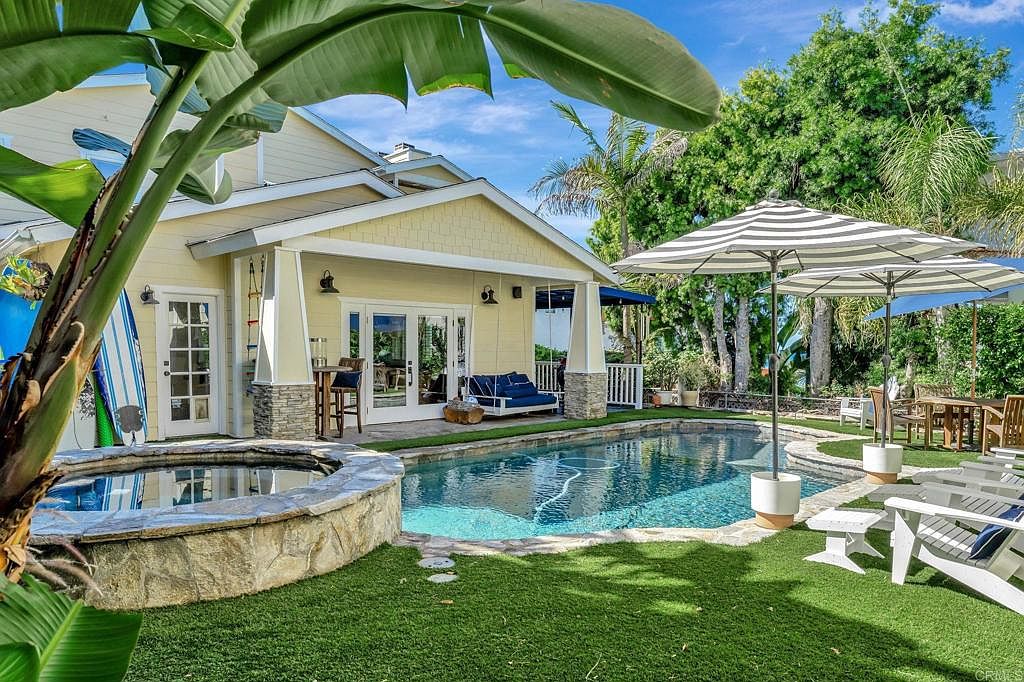 This image showcases a beautiful backyard featuring a pool and spa area. The pool is surrounded by lush green artificial turf and stone accents, with lounge chairs and umbrellas providing a relaxing atmosphere. The house, with its yellow siding and white trim, serves as a backdrop, creating an inviting outdoor living space.
