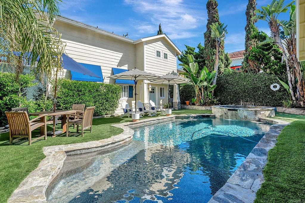 This image showcases a beautifully landscaped backyard featuring a sparkling pool and spa area. The pool is surrounded by lush greenery and stone accents, creating a serene and inviting atmosphere. The house is visible in the background, complemented by striped umbrellas and outdoor furniture, suggesting a perfect space for relaxation and entertainment.