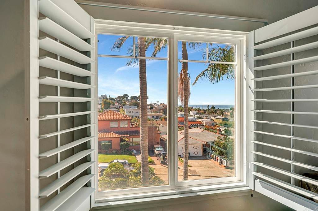 The image showcases a view from inside a room, likely a bedroom, through a double window with white shutters partially open on either side. Outside, palm trees frame a scenic view of a residential neighborhood leading towards the ocean under a clear blue sky. The scene evokes a sense of tranquility and connection to the outdoors.