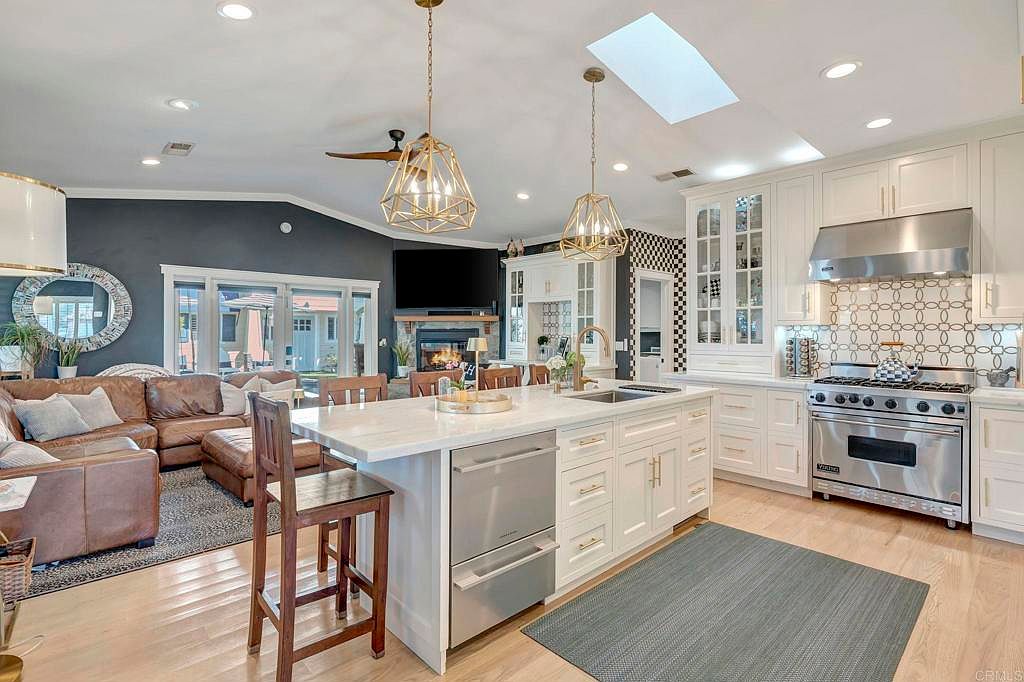 This is a well-lit, open-concept kitchen and living area featuring a large kitchen island with white cabinetry and a marble countertop. Stainless steel appliances and pendant lighting add a modern touch, while a comfortable leather sectional sofa is visible in the adjacent living area. The perspective is from a wide angle, capturing the spaciousness and connection between the kitchen and living room.