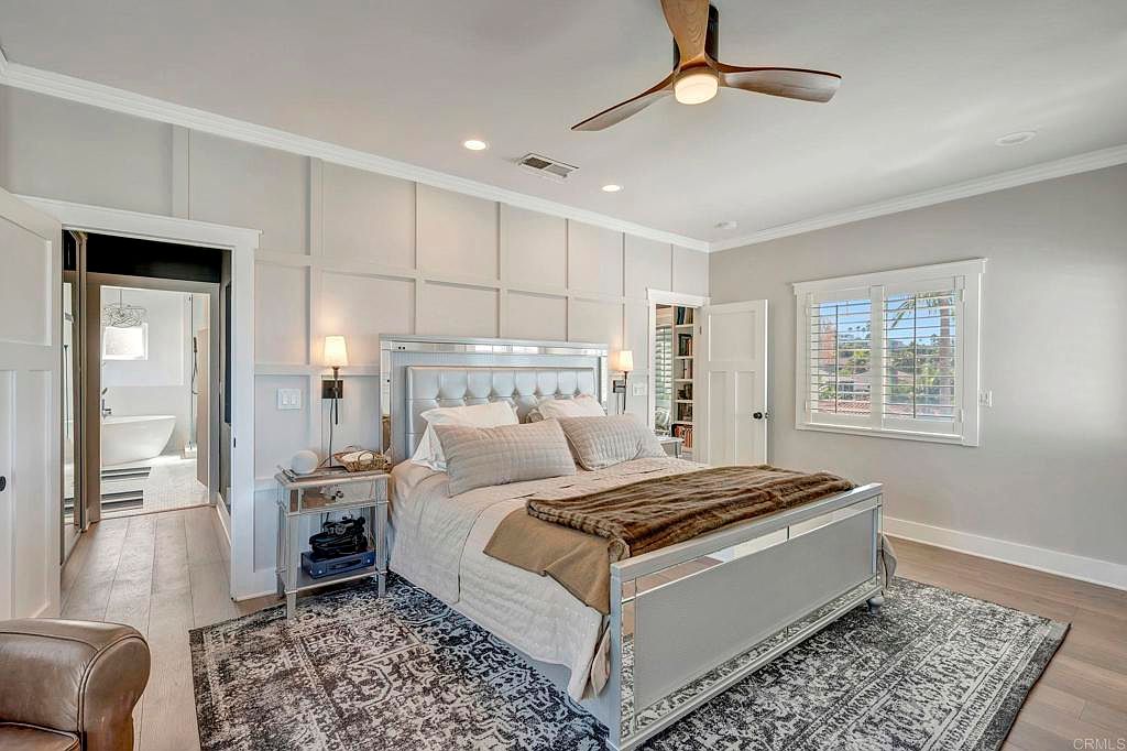 This is a well-lit primary bedroom featuring a mirrored bed frame and matching nightstand, complemented by a neutral-toned rug and bedding. The room has a sophisticated and calming atmosphere, enhanced by the paneled wall detail and a view to an adjacent bathroom. A ceiling fan adds a touch of comfort.